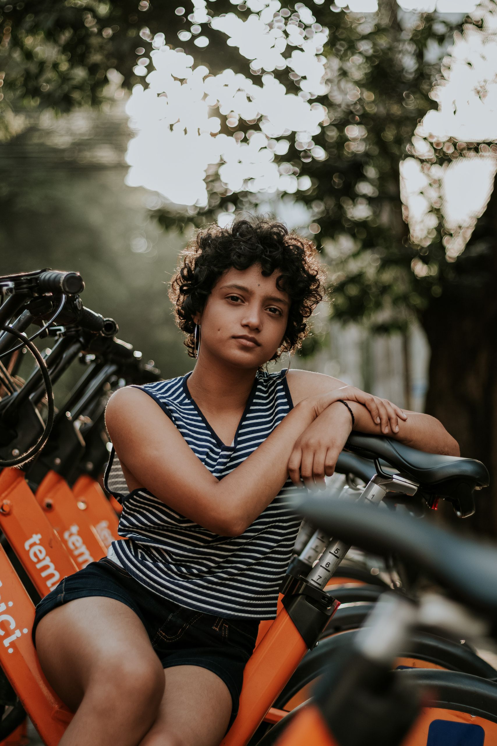 A woman leans casually on a row of orange rental bicycles in an outdoor setting.