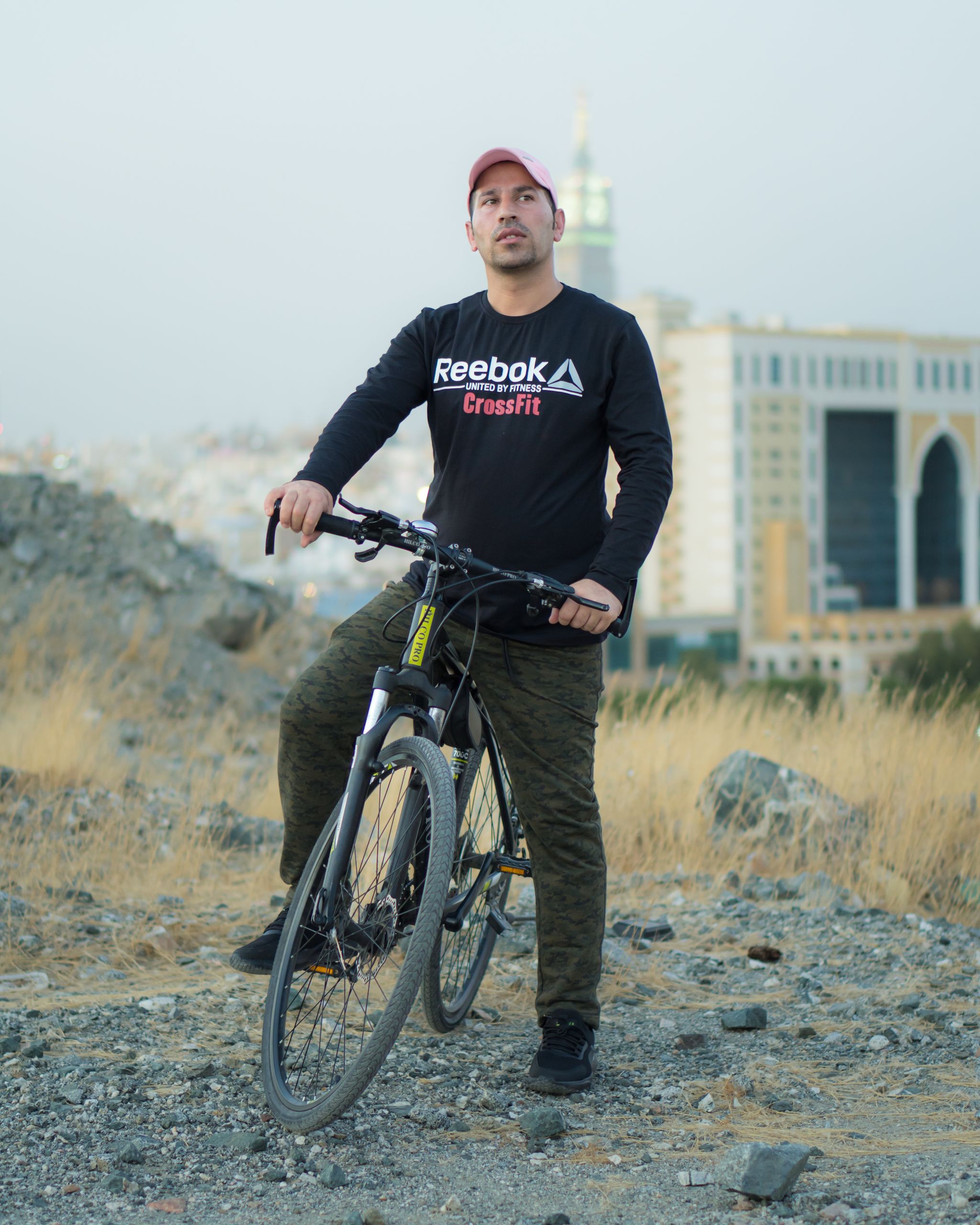 Caucasian man in black sweatshirt and pink cap standing with mountain bike outdoors in cityscape.