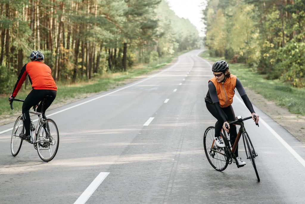 Two cyclists biking on an empty road through a lush forest, wearing sportswear.
