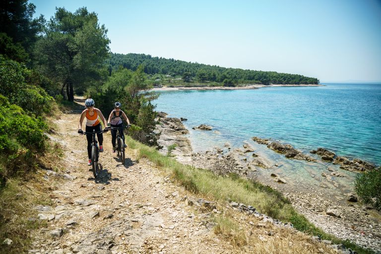 Two cyclists ride on a scenic coastal trail by the sea on a sunny day, showcasing adventure and nature.