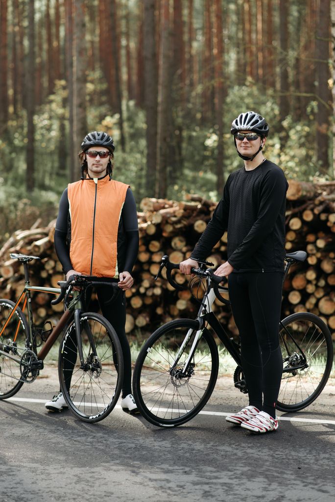 Two men with bicycles standing in front of wood logs in a forest setting, wearing helmets and activewear.